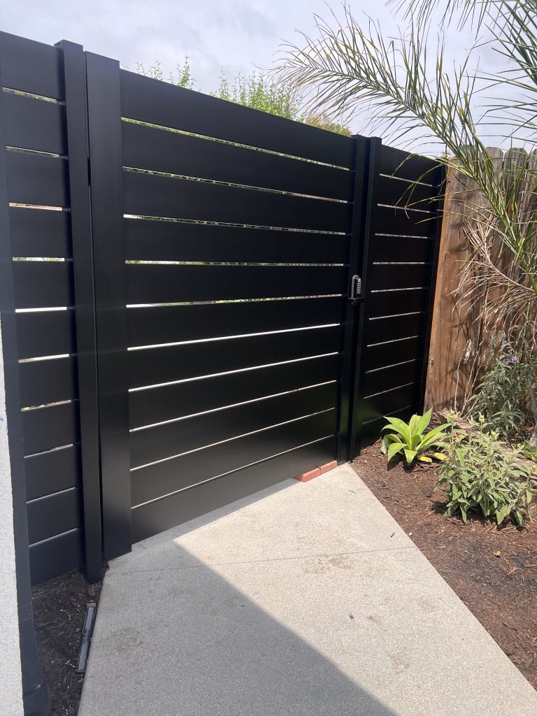 Black aluminum cantilever gate with horizontal slats, showcasing a modern design, set against a landscaped pathway and greenery, emphasizing low-maintenance and decorative features.